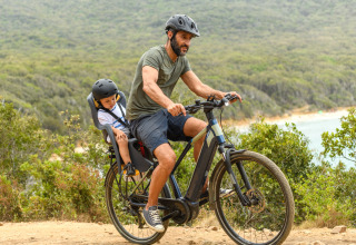 Uomo in bici con bambino nel seggiolino percorre un sentiero naturalistico di un villaggio glamping.