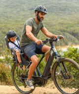 Hombre pedalea con un niño en asiento infantil por un sendero natural en un parque vacacional, con cascos.