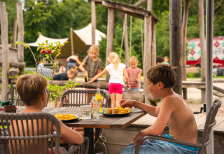 Kinderen genieten van een maaltijd op het terras van vakantiepark Sandberghe, met spelende kinderen op de achtergrond.