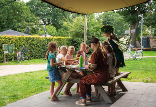 Kinderen knutselen buiten aan een picknicktafel in Sandberghe, Noord-Brabant, begeleid door iemand verkleed als fee.