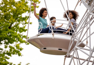 Drie lachende mensen genieten van het reuzenrad in een vakantiepark met glamping, omringd door bomen.