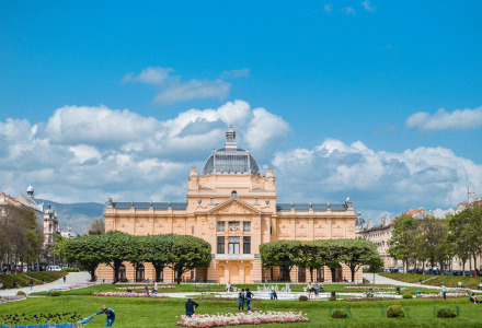 Fachada de edificio histórico con cúpula de cristal, jardines verdes y personas paseando en día soleado.