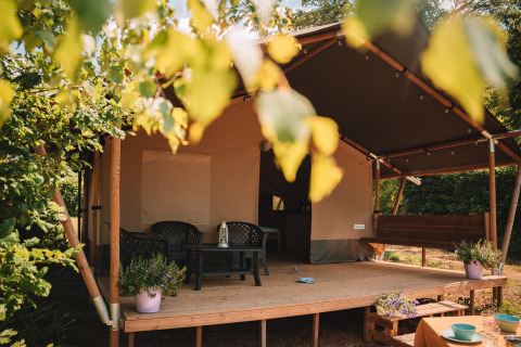 Safari tent with a covered porch, plastic chairs and table, potted plants, surrounded by green trees.