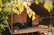 Safari tent with a covered porch, plastic chairs and table, potted plants, surrounded by green trees.