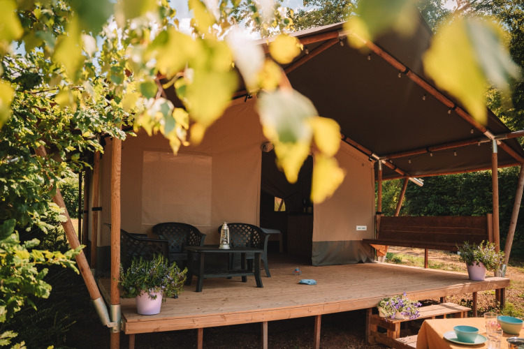 Safari-Zelt mit überdachter Terrasse, Gartenmöbeln und Blumen, umgeben von Bäumen im Sonnenlicht.