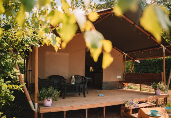 Safari tent with a covered porch, plastic chairs and table, potted plants, surrounded by green trees.