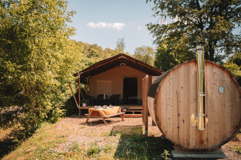 Outdoor photo of a safari tent with wooden sauna and picnic table, surrounded by lush green trees.