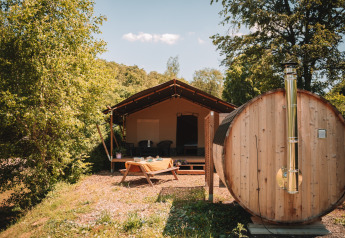Outdoor photo of a safari tent with wooden sauna and picnic table, surrounded by lush green trees.