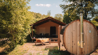 Outdoor photo of a safari tent with wooden sauna and picnic table, surrounded by lush green trees.