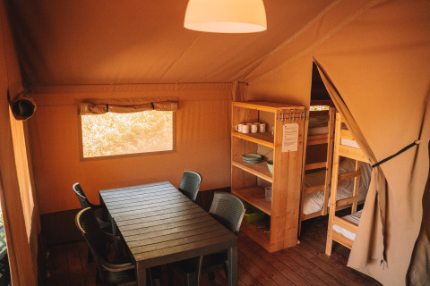 Interior of a safari tent with dining table, chairs, wooden shelves, and bunk beds, cozy rustic atmosphere.