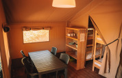 Interior of a safari tent with dining table, chairs, wooden shelves, and bunk beds, cozy rustic atmosphere.