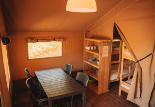Interior of a safari tent with dining table, chairs, wooden shelves, and bunk beds, cozy rustic atmosphere.