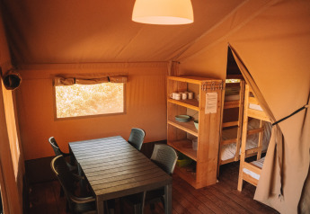 Interno di una tenda safari con tavolo da pranzo, sedie, scaffali in legno e letti a castello, atmosfera accogliente.