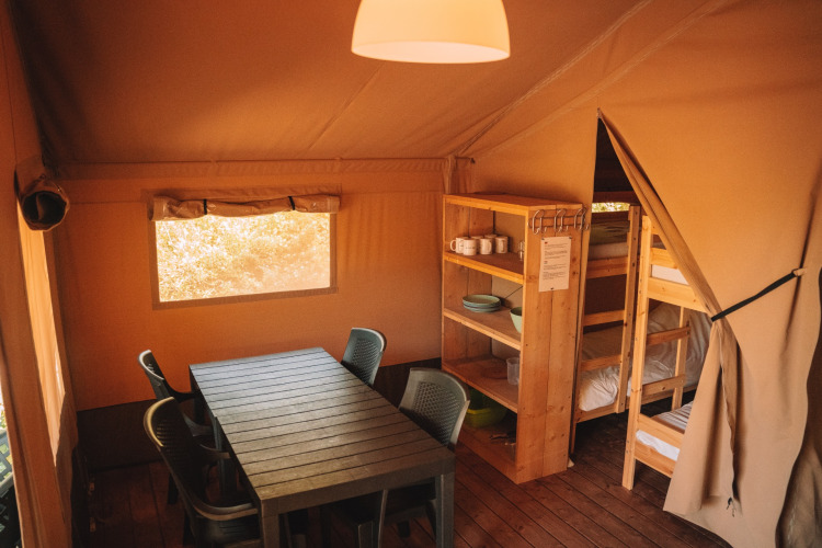 Interior de una tienda safari con mesa, sillas, estantería de madera y literas, ambiente rústico y acogedor.