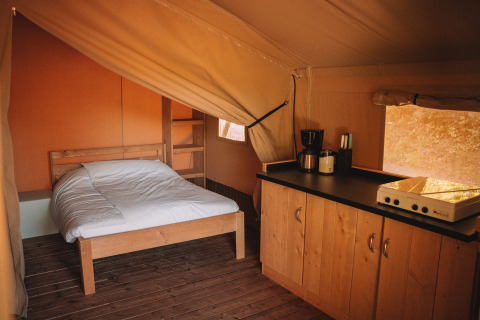 Interior de una tienda safari con cama doble, suelo de madera y cocina con cafetera y utensilios.