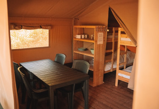 Interieur van een safaritent met een eettafel, stoelen, stapelbedden en open rekken met servies.