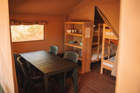 Interior of a safari tent featuring a dining table, chairs, bunk beds, and open shelves with dishes.