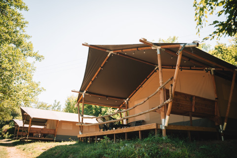 Large safari tents with wooden frames and porches on a scenic campground, surrounded by green trees.