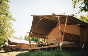 Large safari tents with wooden frames and porches on a scenic campground, surrounded by green trees.