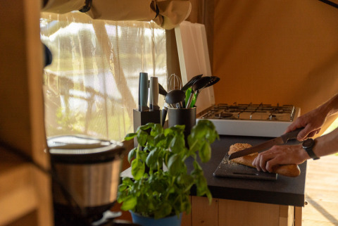 Interior de la tienda safari Maaszicht en Camping de Boomgaard, persona cortando pan en la cocina.