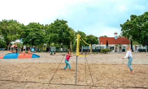 Twee vrouwen spelen beachvolleybal op het zand met vakantiepark Bad Hoophuizen op de achtergrond in Gelderland.