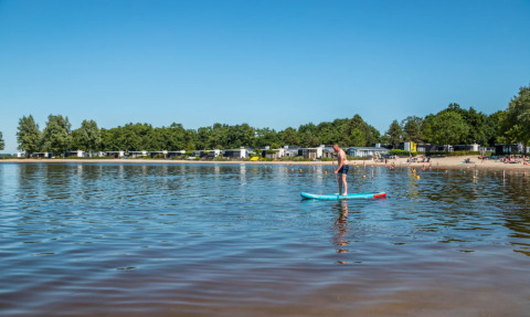 Man doet aan suppen op een kalm meer bij Hulshorst, omgeven door bomen, strand en vakantiehuisjes.