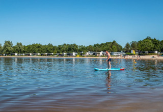 Man doet aan suppen op een kalm meer bij Hulshorst, omgeven door bomen, strand en vakantiehuisjes.