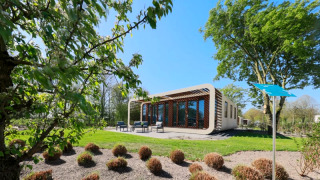 Lodge Modus moderne avec grandes baies vitrées et terrasse, entouré d’arbres sous un ciel bleu clair.