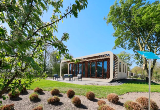 Lodge Modus moderne avec grandes baies vitrées et terrasse, entouré d’arbres sous un ciel bleu clair.