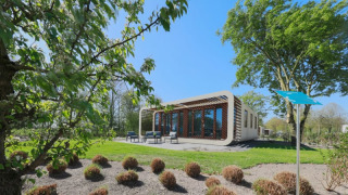 Moderne Modus-lodge met grote ramen en terras, omringd door bomen en struiken onder een blauwe lucht.