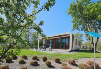 Moderne Modus-lodge med store vinduer og terrasse, omgivet af grønne træer og buske under en blå himmel.