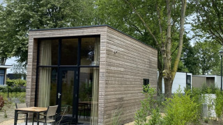 Moderne glamping cabin met houten bekleding, grote ramen en terras, omgeven door bomen en beplanting.