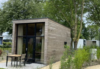 Et moderne glamping-hus med trælister, store vinduer, terrasse og borde, omgivet af grønne træer og planter.