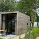Et moderne glamping-hus med trælister, store vinduer, terrasse og borde, omgivet af grønne træer og planter.