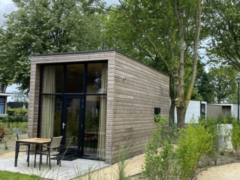 Cabane de glamping moderne en bois, grandes fenêtres et terrasse, entourée d’arbres et de verdure.