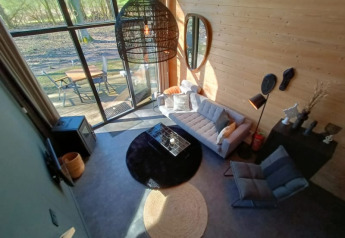 Sfeervolle glamping woonkamer met grote ramen, moderne bank en buitenterras met uitzicht op de natuur.