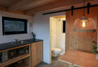 Cocina y comedor minimalista con detalles en madera, lámpara colgante y baño abierto en Tiny House Holanda.