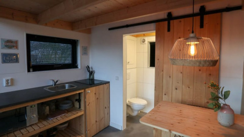 Minimalistisk køkken og spisestue med træindretning, synligt toilet og hyggelig belysning i en Tiny House-lodge.