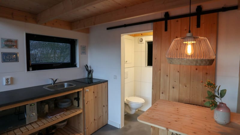 Cocina y comedor minimalista con detalles en madera, lámpara colgante y baño abierto en Tiny House Holanda.