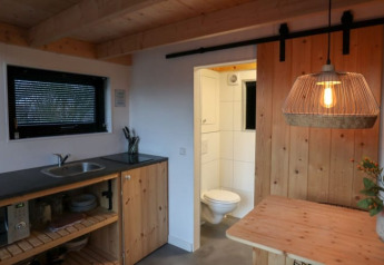 Cocina y comedor minimalista con detalles en madera, lámpara colgante y baño abierto en Tiny House Holanda.