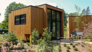 Lodge moderne en bois avec de grandes fenêtres, terrasse et jardin naturel, entouré d’arbres et de plantes.