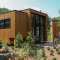 Moderne houten lodge met grote ramen en terras, omgeven door natuurlijke tuin en groene bomen.
