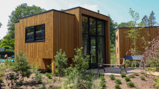 Moderne houten lodge met grote ramen en terras, omgeven door natuurlijke tuin en groene bomen.