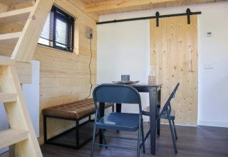 Interno lodge luminoso con pannelli in legno, tavolo, due sedie, panca, porta scorrevole e scala a soppalco.