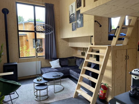 Pequeña casa moderna con entrepiso, sofá gris, interior de madera y gran ventana en Bad Hoophuizen.