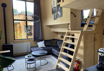 Pequeña casa moderna con entrepiso, sofá gris, interior de madera y gran ventana en Bad Hoophuizen.