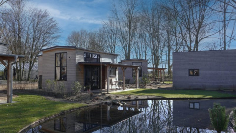 Moderne tiny house in Bad Hoophuizen, Nederland, met een vijver, bomen en een mooie blauwe hemel.