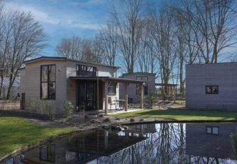 Moderne tiny house in Bad Hoophuizen, Nederland, met een vijver, bomen en een mooie blauwe hemel.