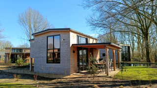 Photo d'une tiny house moderne avec façade en bois et grandes fenêtres, baignée de soleil parmi les arbres.