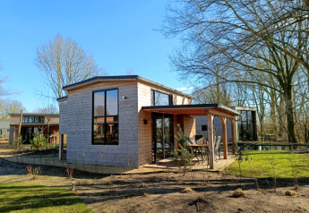 Foto eines modernen Tiny House mit Holzfassade und großen Fenstern in einer sonnigen, grünen Umgebung.